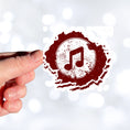 Load image into Gallery viewer, This individual die-cut sticker features a double music note in brown with a paint splattered background. Perfect for performers and music lovers alike!  This image shows a hand holding the music note sticker.
