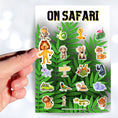 Load image into Gallery viewer, Who's ready to go "On Safari"? This jungle themed sticker sheet has images of your favorite jungle creatures, kid explorers, and all the things you need for your next jungle adventure!  This image shows a hand holding a lion sticker above the On Safari sticker sheet.
