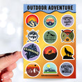 Load image into Gallery viewer, Tie up your boots and enter the wild with this sticker sheet! This sticker sheet has twelve different stickers showing different outdoor activities. From the mountains to camping to climbing this set is perfect for any outdoors person. This image shows a hand holding a sticker showing a climber on a mountain that says "Climbing".

