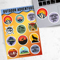 Load image into Gallery viewer, Tie up your boots and enter the wild with this sticker sheet! This sticker sheet has twelve different stickers showing different outdoor activities. From the mountains to camping to climbing this set is perfect for any outdoors person. This image shows the sticker sheet next to an open laptop with stickers of a mountain and one that says "Camping is my happy place"
