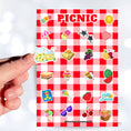 Load image into Gallery viewer, Pack the picnic basket and grab a blanket; we're going on a picnic! This sticker sheet has sticker images of all your favorite picnic foods and activities like sandwiches, fruit, ice cream, and lemonade all on a red and white checked tablecloth background. This image shows a hand holding a sticker of lemonade in a pitcher and glass above the sticker sheet.
