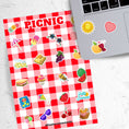 Load image into Gallery viewer, Pack the picnic basket and grab a blanket; we're going on a picnic! This sticker sheet has sticker images of all your favorite picnic foods and activities like sandwiches, fruit, ice cream, and lemonade all on a red and white checked tablecloth background. This image shows the sticker sheet next to an open laptop with stickers of the sun, a red and white checked heart, and an ant holding a yellow apple, applied below the keyboard.
