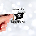 Load image into Gallery viewer, If you think a pirate's life is for you, then this individual die-cut sticker is just what you need! This pirate's life sticker features a jolly rodger (skull and crossed swords) flag with storm clouds and the saying "A Pirate's Life For Me". This image shows a hand holding the pirate's life sticker.
