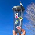 Load image into Gallery viewer, This sticker sheet has 20 different sticker images of fairies and it features a holographic sparkle overlay to give it that magical feel. This image shows a water bottle with stickers of a fairy standing on a pink rose, a fairy on a swing below a big tree, a fairy with a four leaf clover, and a fairy leaning on her elbows with her feet crossed, applied to it.
