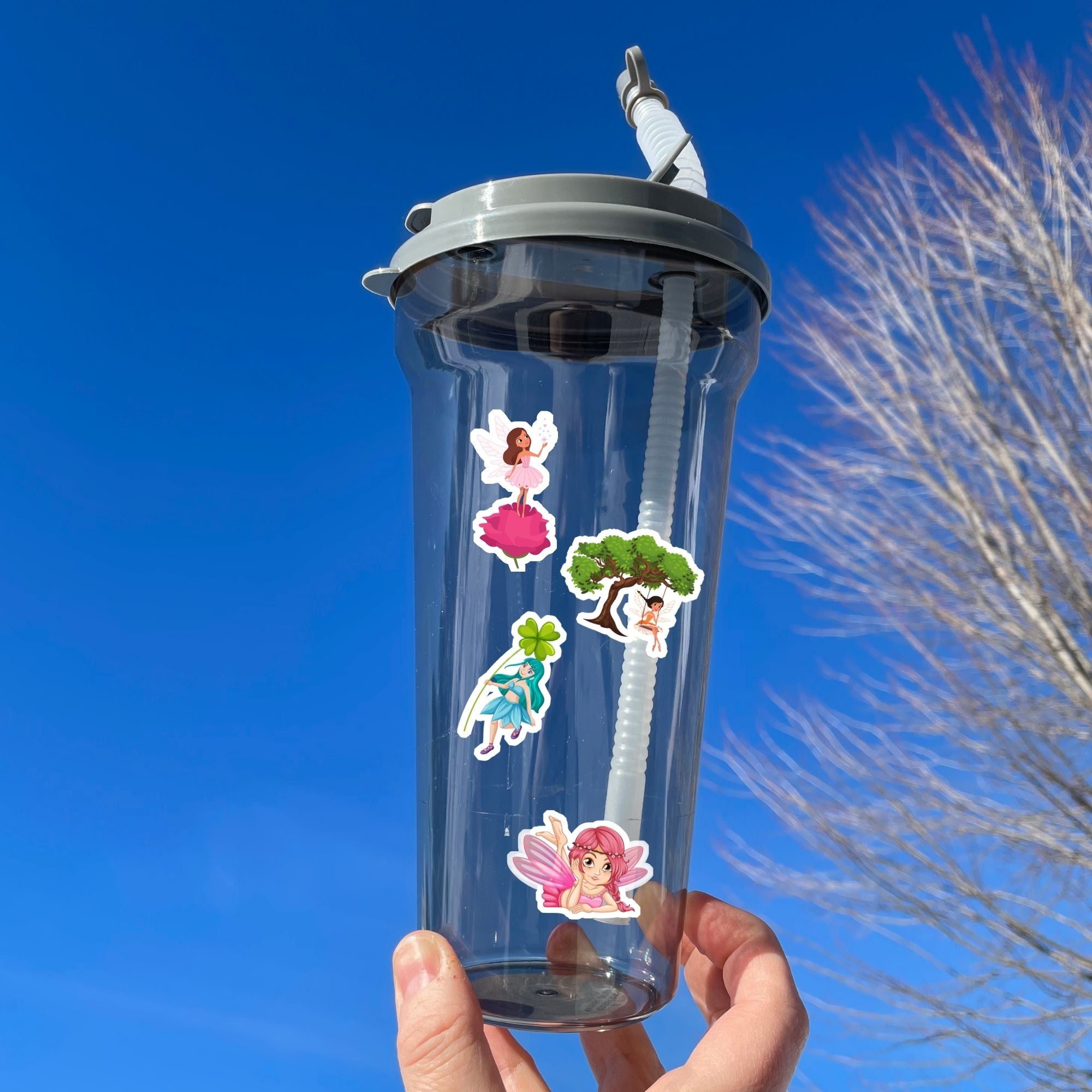 This sticker sheet has 20 different sticker images of fairies and it features a holographic sparkle overlay to give it that magical feel. This image shows a water bottle with stickers of a fairy standing on a pink rose, a fairy on a swing below a big tree, a fairy with a four leaf clover, and a fairy leaning on her elbows with her feet crossed, applied to it.
