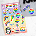 Load image into Gallery viewer, Show your LGBTQ Pride with this sticker sheet! This sticker sheet is filled with rainbow Pride images that are perfect for anywhere you want to show your Pride. This image is of the sticker sheet next to an open laptop with a sticker of heart rainbow glasses and an sticker saying Just Love applied below the keyboard.
