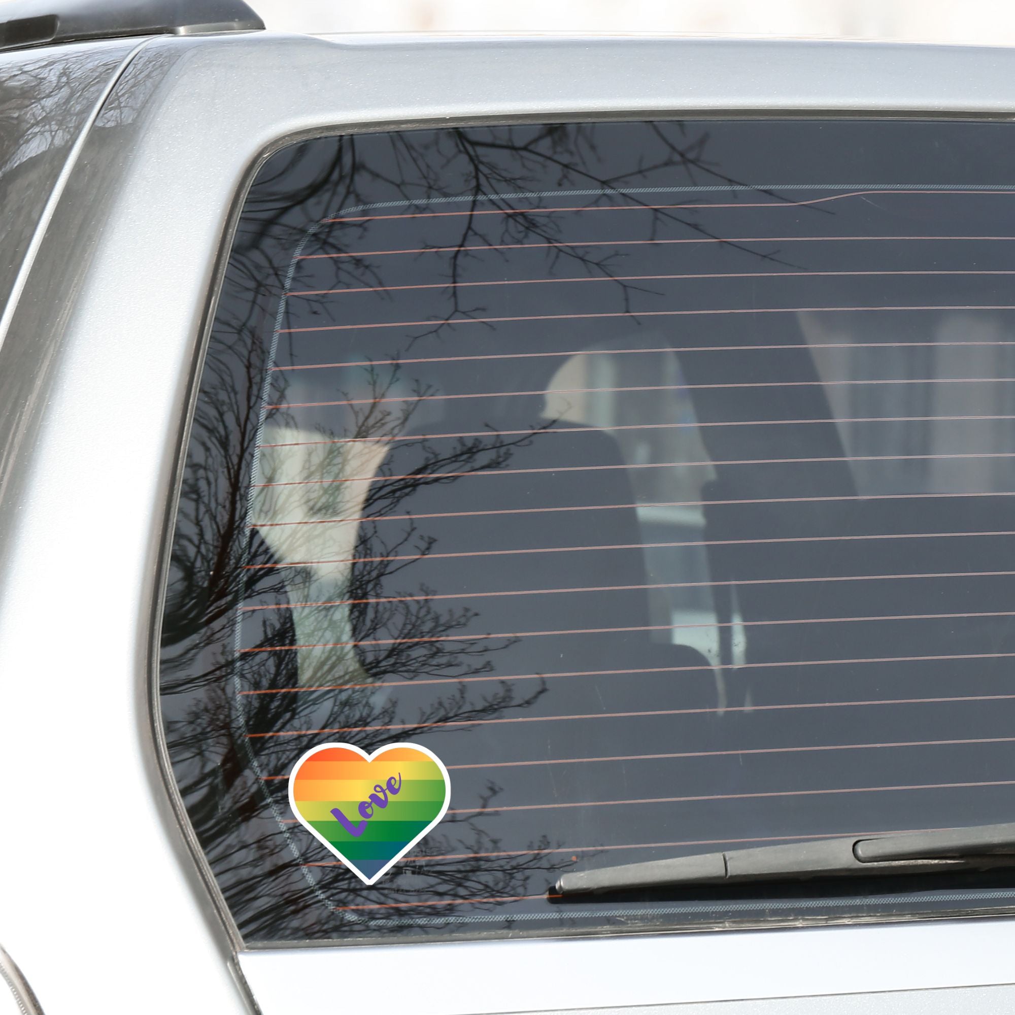 This individual die-cut sticker features a heart shape with graduated colors from orange at the top to blue at the bottom, and the word love written across. Perfect for the people you love! This image shows the Love Heart sticker on the back window of a car.