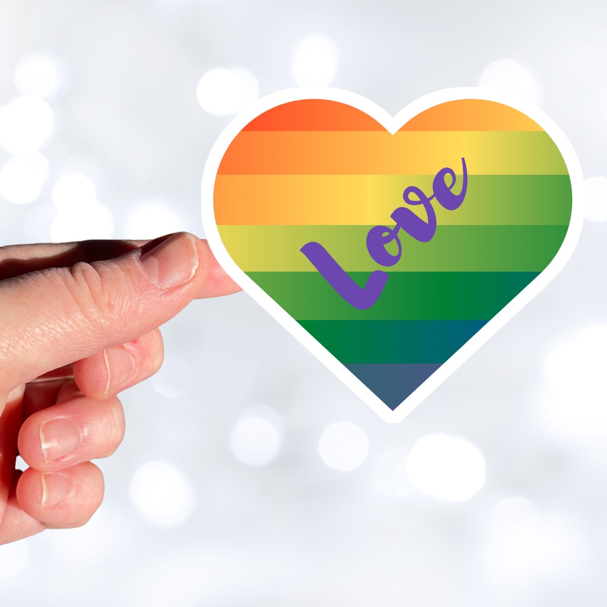 This individual die-cut sticker features a heart shape with graduated colors from orange at the top to blue at the bottom, and the word love written across. Perfect for the people you love! This image shows a hand holding the Love Heart sticker.