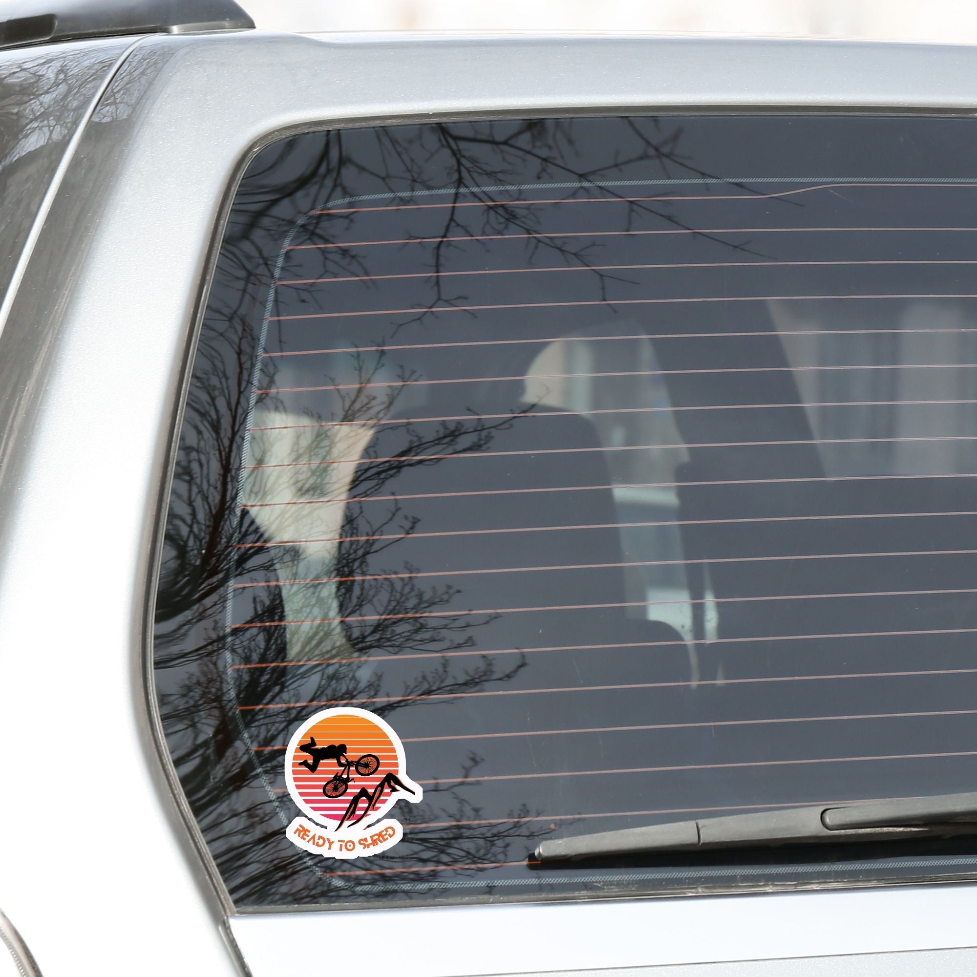 Grab your off-road bicycle and hit the trails! This individual die-cut sticker features the silhouette of a mountain biker flying above mountains, on an orange and white gradient background, with the words "Ready to Shred" below. This image shows the mountain bike sticker on the rear window of a car.