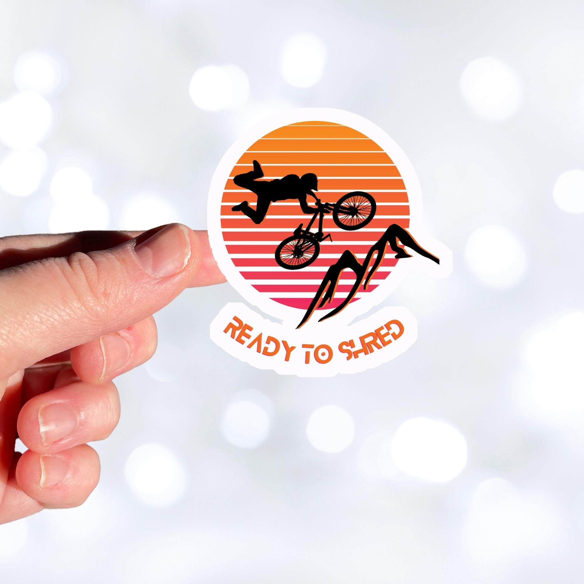 Grab your off-road bicycle and hit the trails! This individual die-cut sticker features the silhouette of a mountain biker flying above mountains, on an orange and white gradient background, with the words "Ready to Shred" below. This image shows a hand holding the mountain bike sticker.