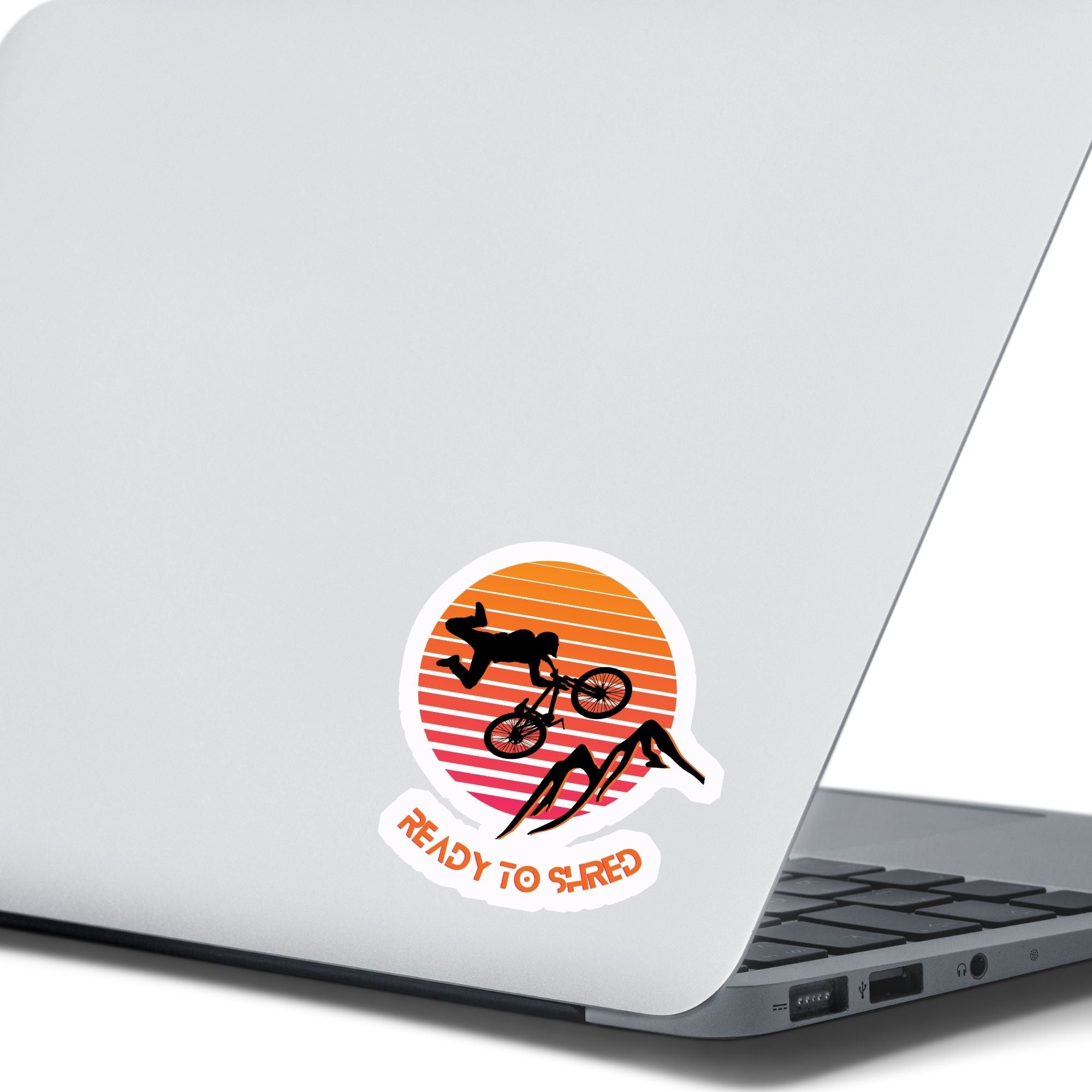 Grab your off-road bicycle and hit the trails! This individual die-cut sticker features the silhouette of a mountain biker flying above mountains, on an orange and white gradient background, with the words "Ready to Shred" below. This image shows the mountain bike sticker on the back of an open laptop.