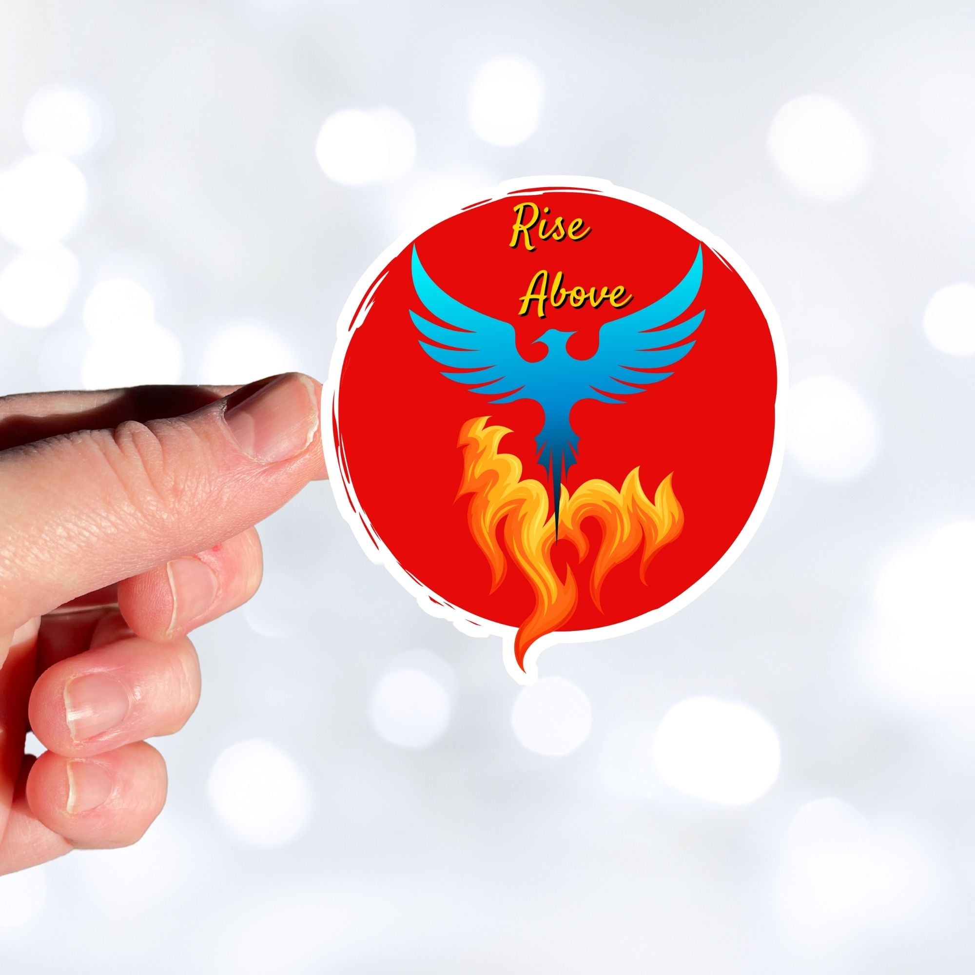 Phoenix rising - this individual die-cut sticker features a blue phoenix rising out of flames with the words "Rise Above" at the top, all on a red background. This image shows a hand holding the Rise Above sticker.