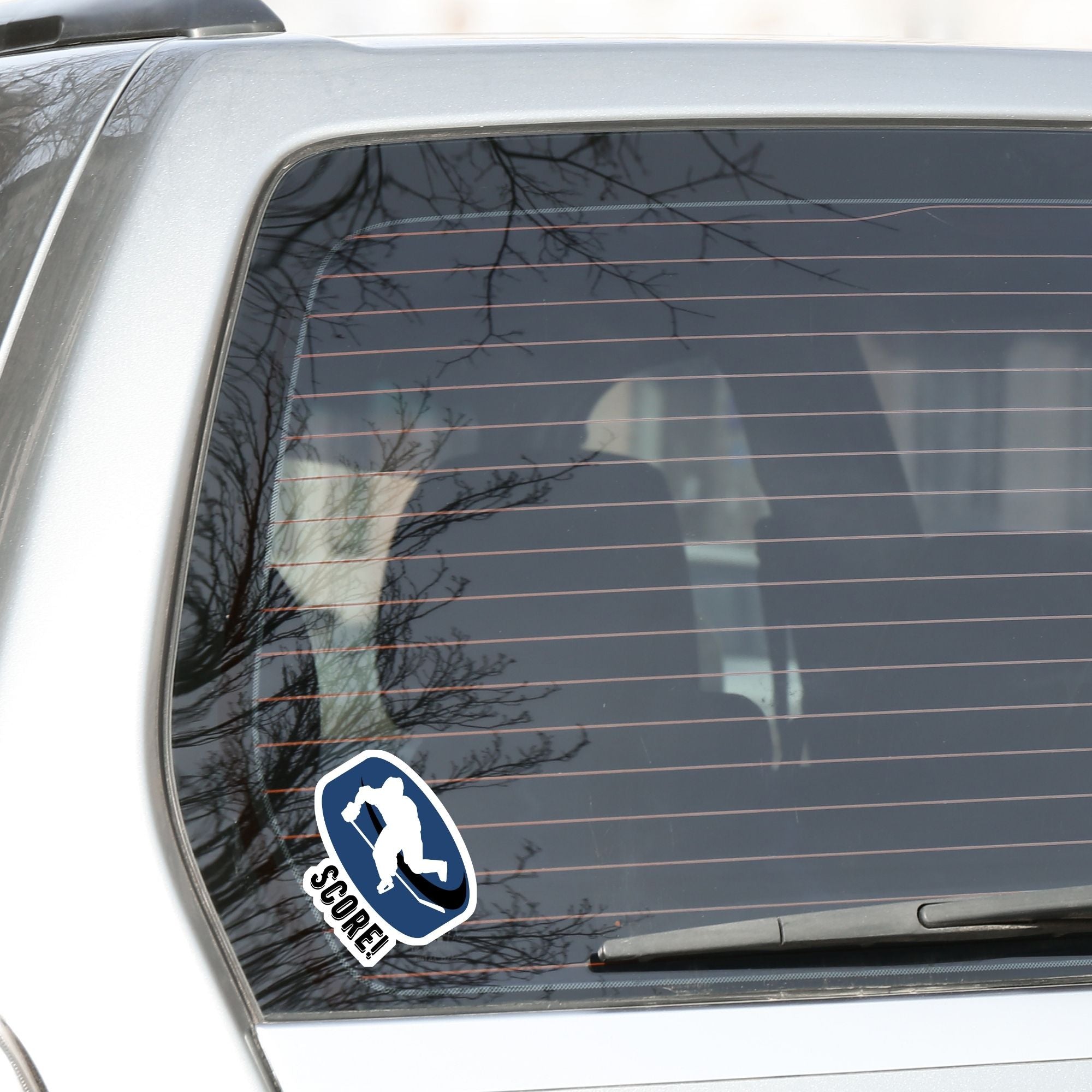 Grab your stick, lace up your skates, and head to the rink! This individual die-cut sticker features a hockey player silhouette on a blue hockey puck with the word "Score!" on the lower left side. This image shows the hockey sticker on the back window of a car.