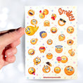 Load image into Gallery viewer, This sticker sheet features all your favorite smiley emojis! It's enough to make anyone smile! This image shows a hand holding a smiley face sticker above the sticker sheet.
