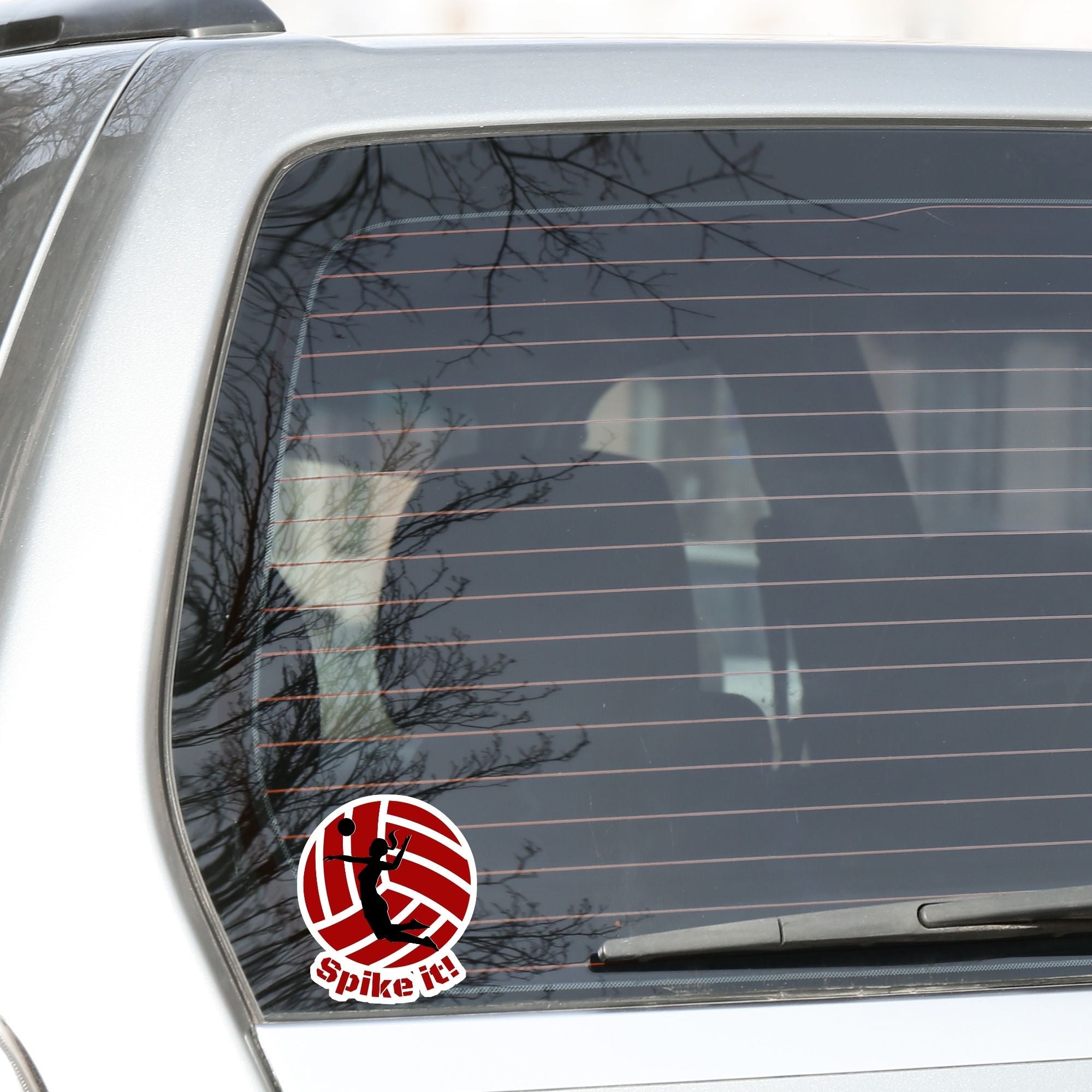 Show your love of volleyball with this individual die-cut sticker! This sticker shows the silhouette of a player with a ponytail about to spike the ball, on a maroon and white volleyball background, with the words "Spike it!" below. This image shows the volleyball sticker on the back window of a car.