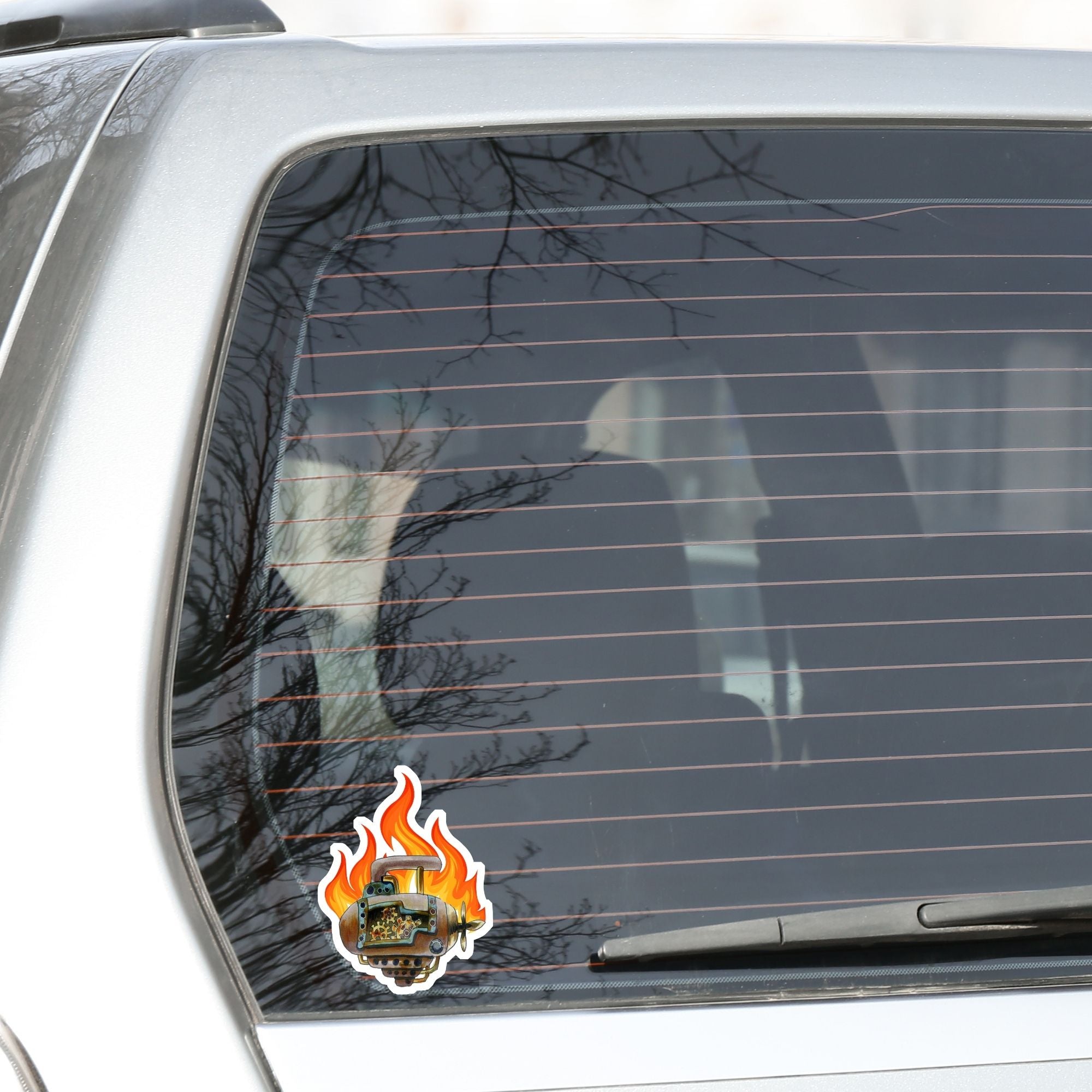 This individual die-cut steampunk sticker features a brown engine with a cutaway to show the gears inside, on a background of yellow, orange, and red flames. This image shows the steampunk engine sticker on the back window of a car.