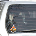 Load image into Gallery viewer, This individual die-cut steampunk sticker features a brown engine with a cutaway to show the gears inside, on a background of yellow, orange, and red flames. This image shows the steampunk engine sticker on the back window of a car.
