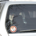 Load image into Gallery viewer, This round individual die-cut sticker features a rusty ring with screws on a white background with two mechanical geckos, one silver and one rust colored, in a yin-yang configuration. This image shows the steampunk geckos sticker on the back window of a car.
