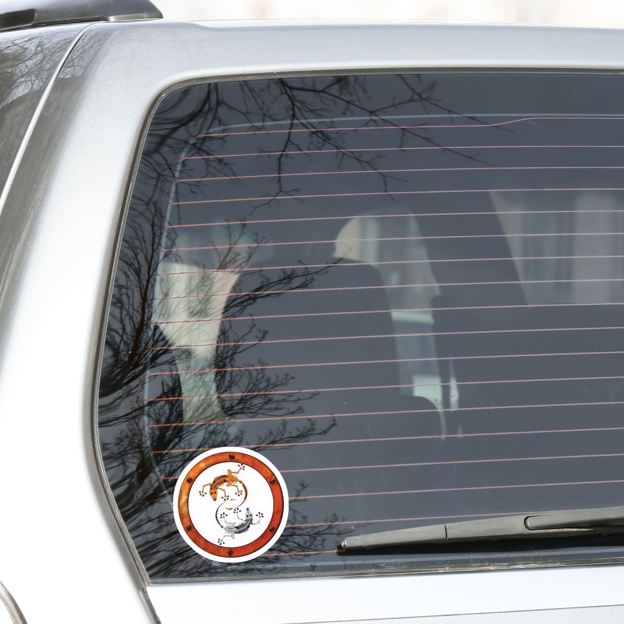 This round individual die-cut sticker features a rusty ring with screws on a white background with two mechanical geckos, one silver and one rust colored, in a yin-yang configuration. This image shows the steampunk geckos sticker on the back window of a car.