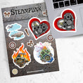 Load image into Gallery viewer, This Steampunk sticker sheet features four large mechanical images: Steampunk Flyers, Steampunk Heart, Steampunk Engine with flames, and Steampunk Engine in pastel, along with smaller miscellaneous gear, butterfly, and pen stickers.  This images shows the sticker sheet next to an open laptop with the Steampunk Heart sticker applied below the keyboard.

