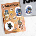 Load image into Gallery viewer, This Steampunk sticker sheet features four large animal stickers: Mr. and Mrs. Owl, Steampunk Monkey, and Steampunk Rotty (Rottweiler) along with smaller miscellaneous gear, butterfly, and pen stickers.  This image shows the sticker sheet next to an open laptop with the Steampunk Rotty sticker applied below the keyboard. The steampunk Rotty sticker is a Rottweiler  wearing a steampunk hat with goggles and a pastel background..
