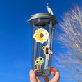 Load image into Gallery viewer, Love sunflowers? Then show it with this sticker sheet! This sticker sheet has nine larger sunflower images. This image shows a water bottle with three stickers - a sunflower morphing into hearts, a woman holding sunflowers, and a boot with sunflowers growing out of it.
