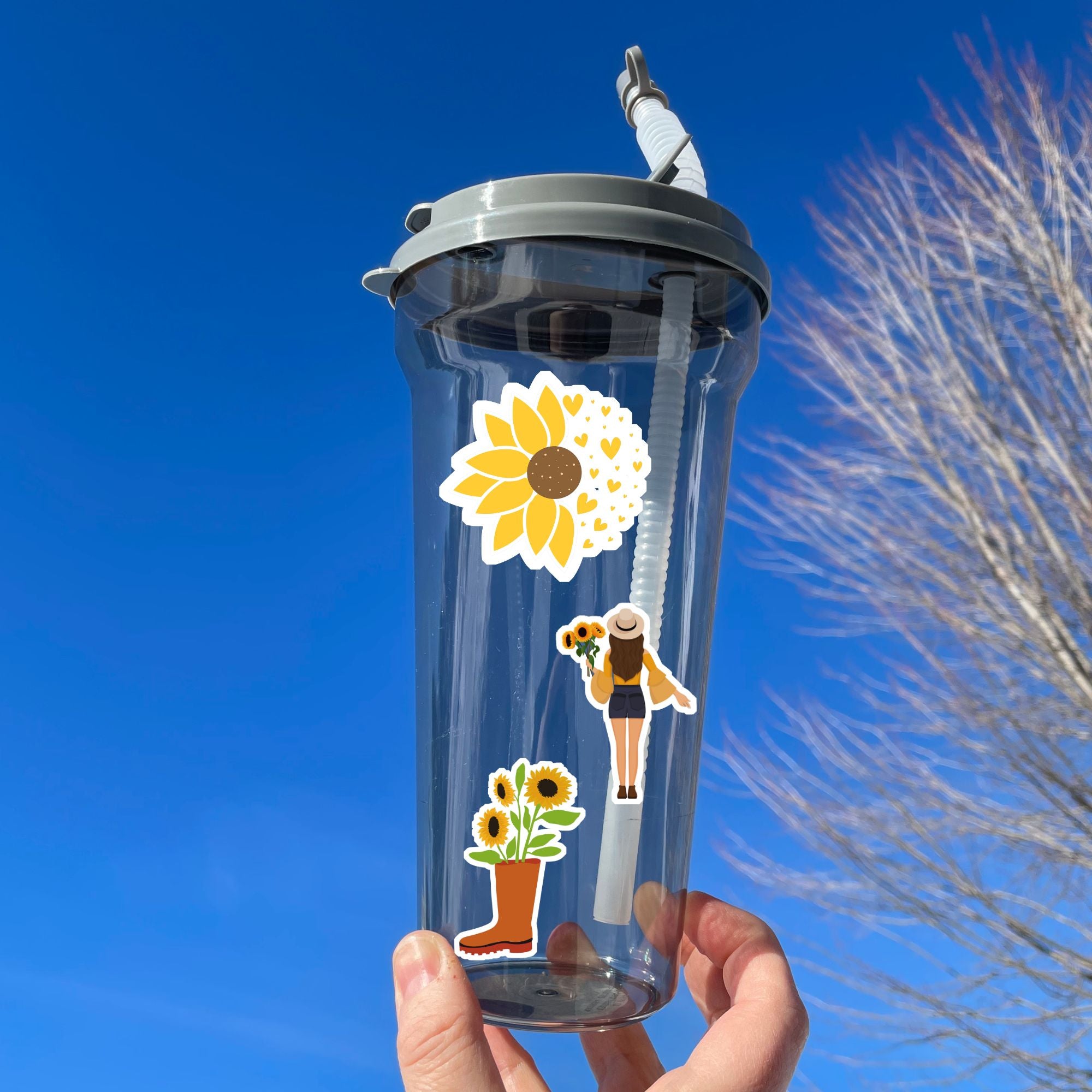 Love sunflowers? Then show it with this sticker sheet! This sticker sheet has nine larger sunflower images. This image shows a water bottle with three stickers - a sunflower morphing into hearts, a woman holding sunflowers, and a boot with sunflowers growing out of it.