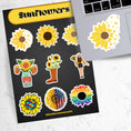 Load image into Gallery viewer, Love sunflowers? Then show it with this sticker sheet! This sticker sheet has nine larger sunflower images. This image shows the sticker sheet next to an open laptop with a sticker of a sunflower morphing into butterflies applied below the keyboard.
