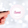 Load image into Gallery viewer, This round individual die-cut sticker is Sweet! It features the word Sweet in the center surrounded by pink and purple circular dots. This makes a great gift for anyone you're sweet on! This image shows a hand holding the Sweet sticker.
