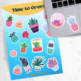 Load image into Gallery viewer, Let your green thumb shine with this sticker sheet. It features a variety of succulent plant stickers that are perfect for your favorite gardener! This image shows the sticker sheet next to an open laptop with a round terrarium sticker and a potted succulent plant sticker, both in pastel colors, applied below the keyboard..
