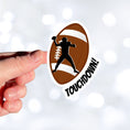 Load image into Gallery viewer, This individual die-cut sticker features the silhouette of a football (American) player about to pass the ball, on a football background, with the word "Touchdown!" on the lower right side. This image shows a hand holding the touchdown sticker.
