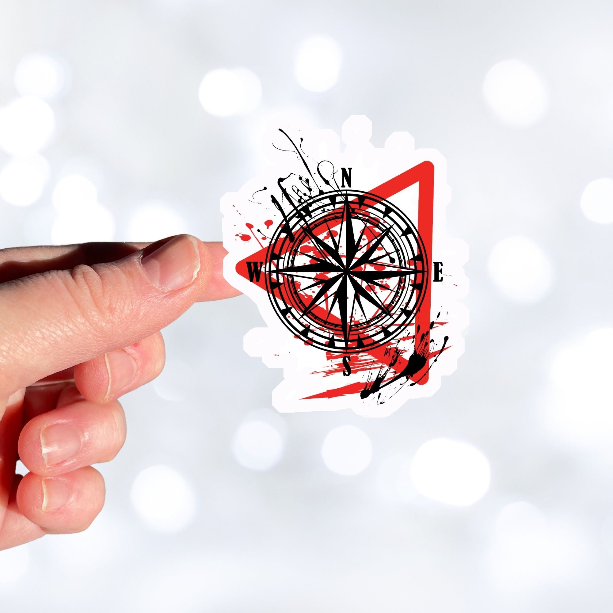 Trash Polka uses red, black, and white with a combination of abstract, surrealistic, and realistic images, and this individual die-cut sticker features a compass inside a red triangle outline on a white background. This image shows a hand holding the trash polka compass sticker.