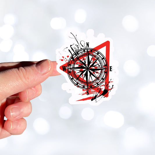 Trash Polka uses red, black, and white with a combination of abstract, surrealistic, and realistic images, and this individual die-cut sticker features a compass inside a red triangle outline on a white background. This image shows a hand holding the trash polka compass sticker.