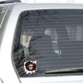 Load image into Gallery viewer, Trash Polka uses red, black, and white with a combination of abstract, surrealistic, and realistic images, and this individual die-cut sticker features a sugar skull woman with roses in her hair on a black background with a red grid. This image shows the trash polka sugar skull sticker on the back window of a car.
