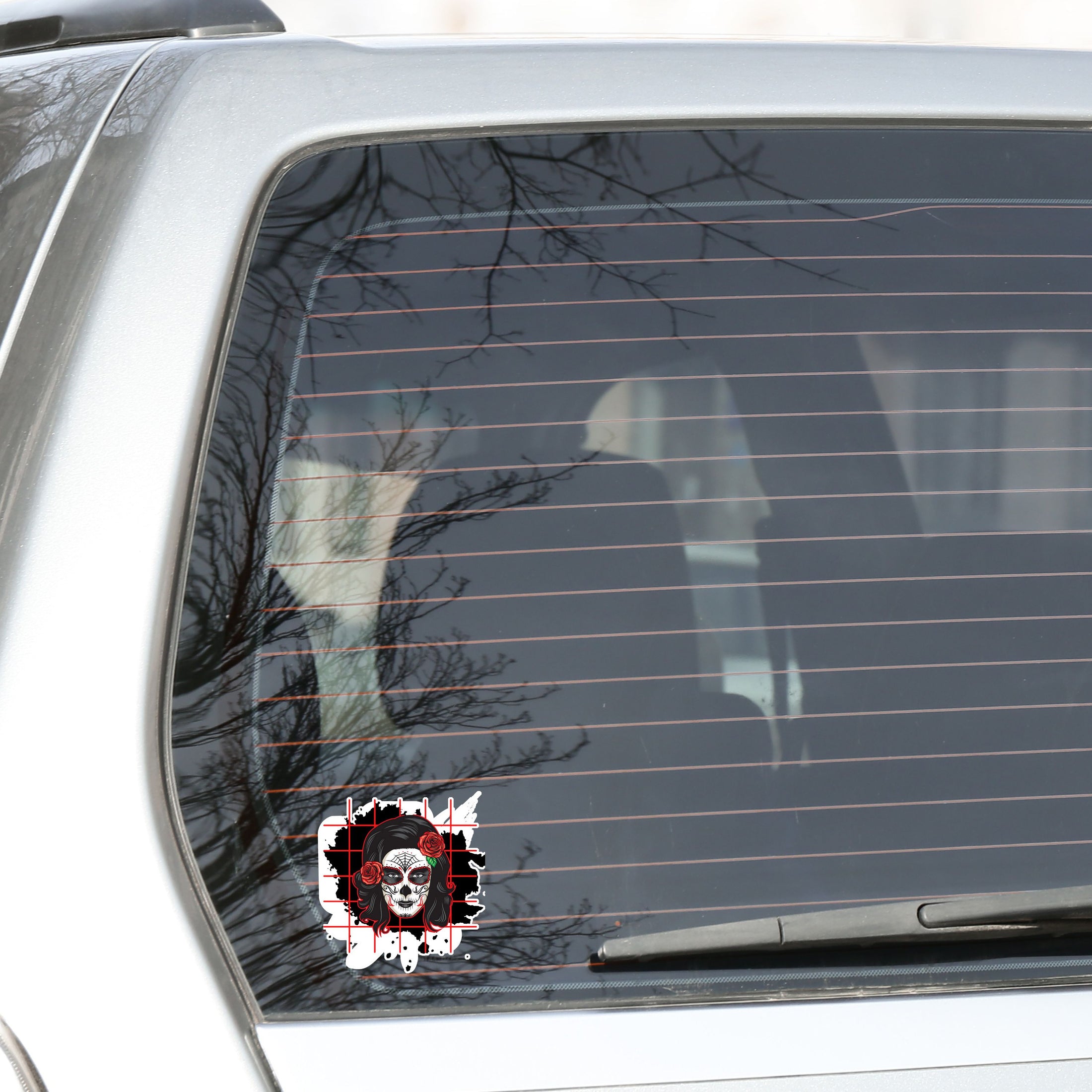 Trash Polka uses red, black, and white with a combination of abstract, surrealistic, and realistic images, and this individual die-cut sticker features a sugar skull woman with roses in her hair on a black background with a red grid. This image shows the trash polka sugar skull sticker on the back window of a car.
