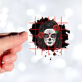 Load image into Gallery viewer, Trash Polka uses red, black, and white with a combination of abstract, surrealistic, and realistic images, and this individual die-cut sticker features a sugar skull woman with roses in her hair on a black background with a red grid. This image shows a hand holding the trash polka sugar skull sticker.
