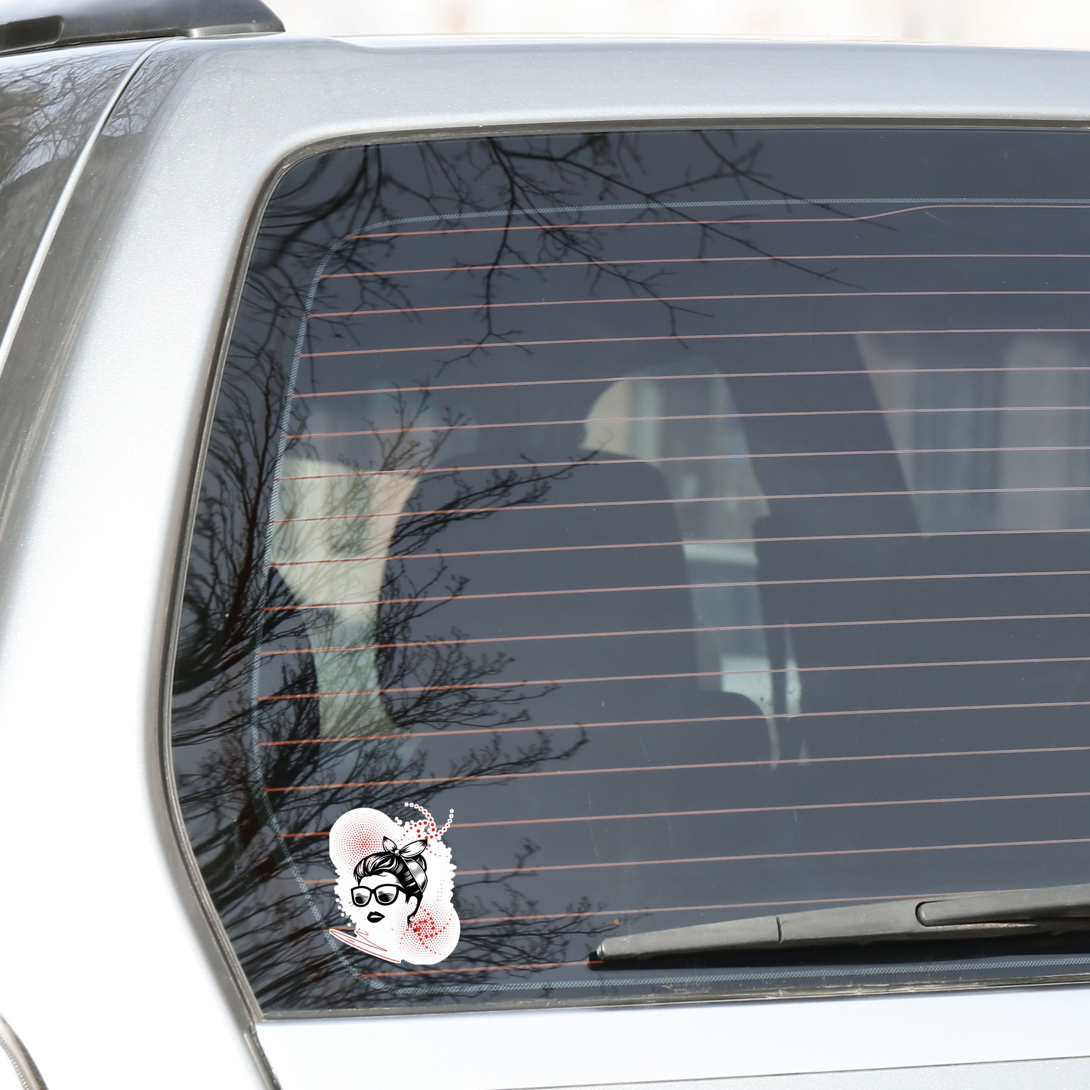 Trash Polka uses red, black, and white with a combination of abstract, surrealistic, and realistic images, and this individual die-cut sticker features a woman wearing sunglasses with her hair tied up on a white background with red swirls. This image shows the trash polka woman sticker on the back window of a car.