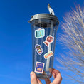 Load image into Gallery viewer, Pack your bags and grab your passport; it's travel time! This sticker sheet is filled with travel related sticker images sure to please even the most discerning traveler. Some of the sticker images are an airplane, suitcase, globe, and ship.  This image is of a water bottle with stickers of a suitcase, a globe, a camera, a bus, and a travel stamp applied to it.
