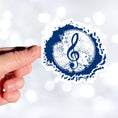 Load image into Gallery viewer, This individual die-cut sticker features a blue treble clef with a paint splattered background. Perfect for performers and music lovers alike!  This image shows a hand holding the treble clef sticker.
