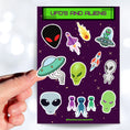 Load image into Gallery viewer, This sticker sheet is filled with aliens and UFOs with individual stickers of aliens, space ships, and alien creatures. This image is of a hand holding one of the individual stickers above the sticker sheet
