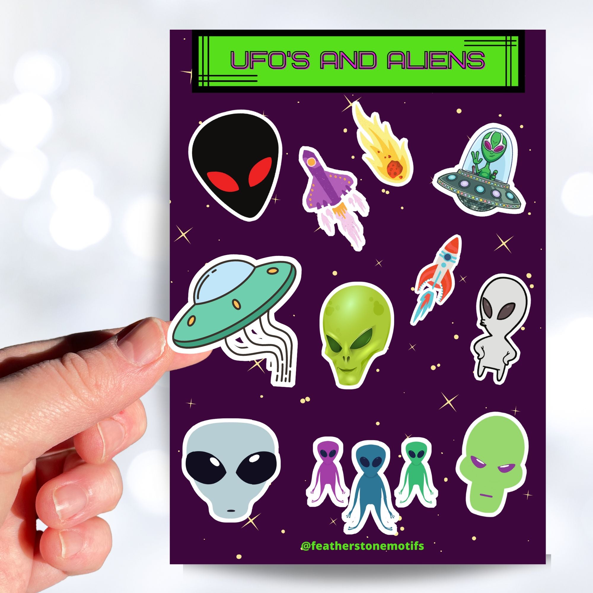 This sticker sheet is filled with aliens and UFOs with individual stickers of aliens, space ships, and alien creatures. This image is of a hand holding one of the individual stickers above the sticker sheet