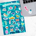 Load image into Gallery viewer, This sticker sheet is filled with fun and cute sticker images of all your favorite undersea creatures! Images include whales, sharks, jellyfish, manatee's, and crabs. There's even a yellow submarine! This image shows a pink seahorse sticker, and a blue jellyfish sticker, applied below the keyboard.
