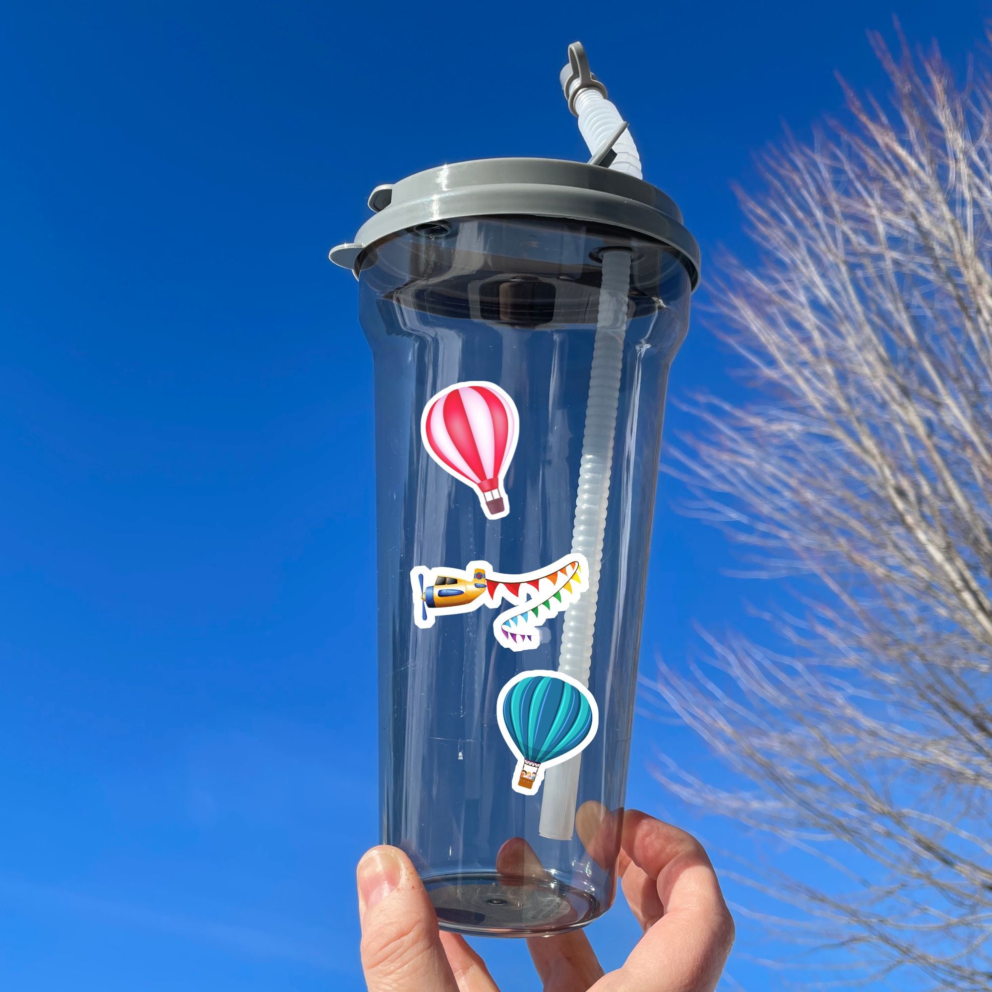 Fly away in a hot air balloon, a plane, or a helicopter with this sticker sheet. This image shows a water bottle with stickers of a red and white hot air balloon, a plan pulling a banner of multi-colored triangle flags, and a blue striped hot air balloon, applied to it.
