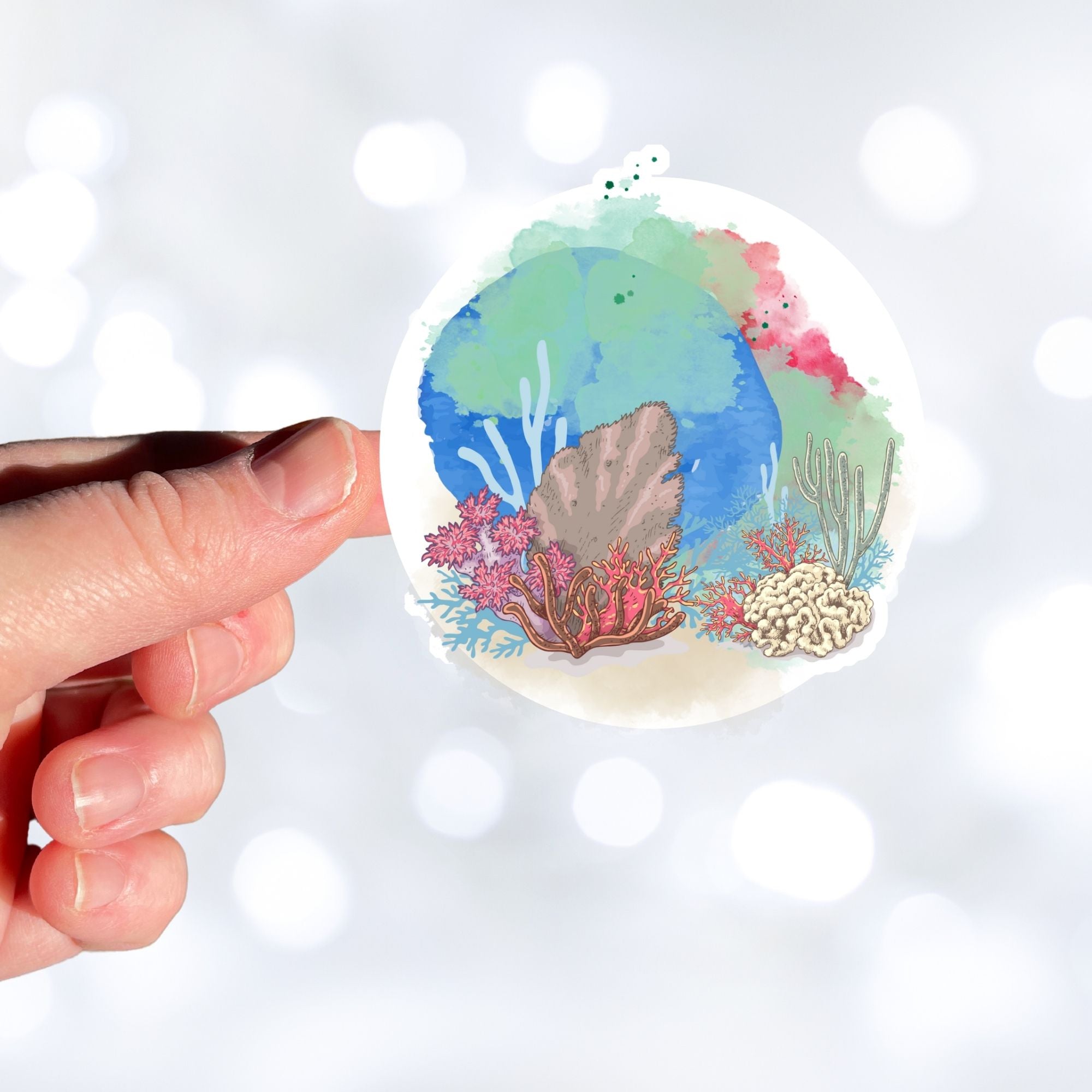 This underwater scene features corals, sand, and a pastel watercolor background on an individual die-cut sticker. With this sticker you can take the reef with you anywhere you go! This image shows a hand holding the Watercolor Reef sticker.