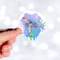 Load image into Gallery viewer, This individual die-cut sticker has a serene looking green sea turtle on a blue and purple background with splash of green. Just looking at this image is relaxing! This image shows a hand holding the Watercolor Sea Turtle sticker.

