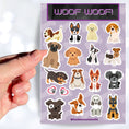 Load image into Gallery viewer, Dog lovers, this sticker sheet is for you! It has sticker images of 16 different dogs plus 2 heart paw stickers. Is your favorite puppy here?  This image shows a hand holding a sticker of a Great Dane puppy above the sticker sheet. 
