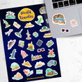Load image into Gallery viewer, Our World Traveler sticker sheet has sticker images of iconic travel destinations! This sheet has a black background with over 30 different stickers including the Statue of Liberty, Easter Island, Stonehenge, and a passport and camera. This image shows the sticker sheet next to an open laptop with stickers of the Grand Canyon, sunglasses, and a travel notebook and pen, applied below the keyboard.
