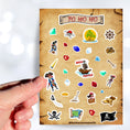 Load image into Gallery viewer, Yo Ho Ho, a Pirate's Life for Me! This sticker sheet is a treasure-worthy find for any pirate with images of the Jolly Roger, treasure, maps, pirates, and a pirate ship. This image is of a hand holding a sticker of a boy pirate above the sticker sheet.
