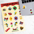Load image into Gallery viewer, This sticker sheet has a sparkle overlay with 19 different stickers of cute ladybugs and bees. This image shows the sticker sheet next to an open laptop with a sticker of a bee mom with her baby bee and a sticker of a ladybug on a flower applied below the keyboard.
