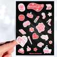 Load image into Gallery viewer, Let's celebrate mom! Mom's can do it all, and this pink aesthetic sticker sheet has sticker images of things that moms, and non-moms, enjoy like - candles, bubble baths, makeup, or coffee. This image shows a hand holding a sticker that says "Mom Life" in the shape of a heart above the sticker sheet.
